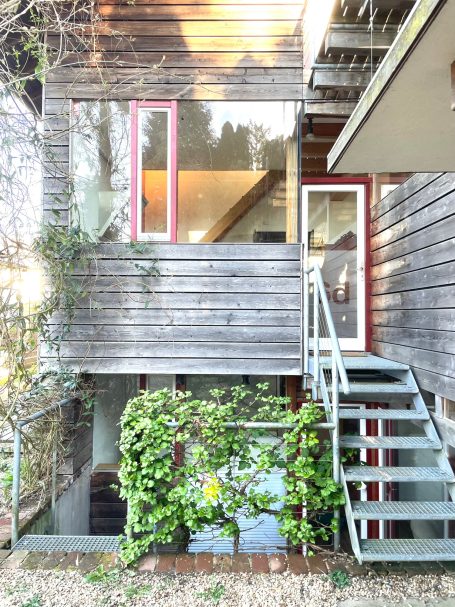 Eingang zu Rumi 4 Holzhütte mit roten Fenstern, Treppe und umgebender grüner Vegetation.